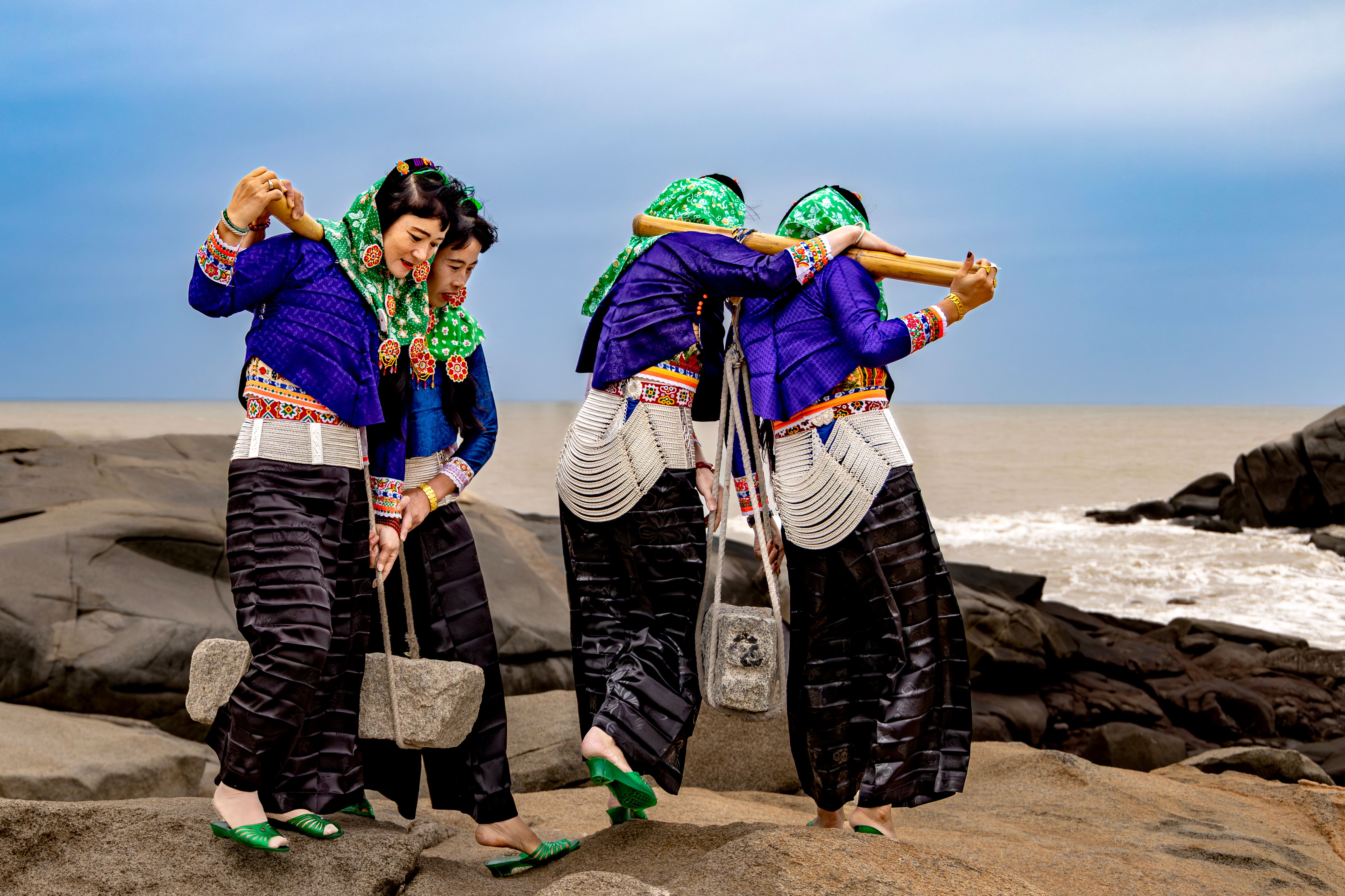 Women in traditional attire perform a cultural dance on a rocky seashore, showcasing vibrant colors.