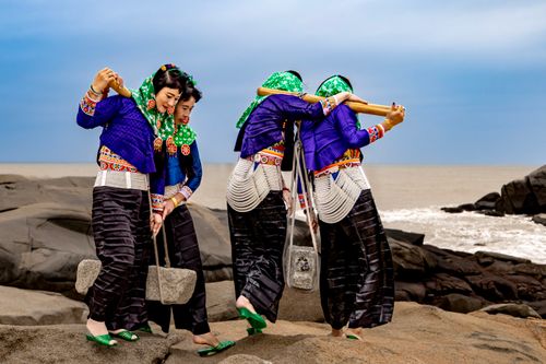 Free Women in traditional attire perform a cultural dance on a rocky seashore, showcasing vibrant colors. Stock Photo