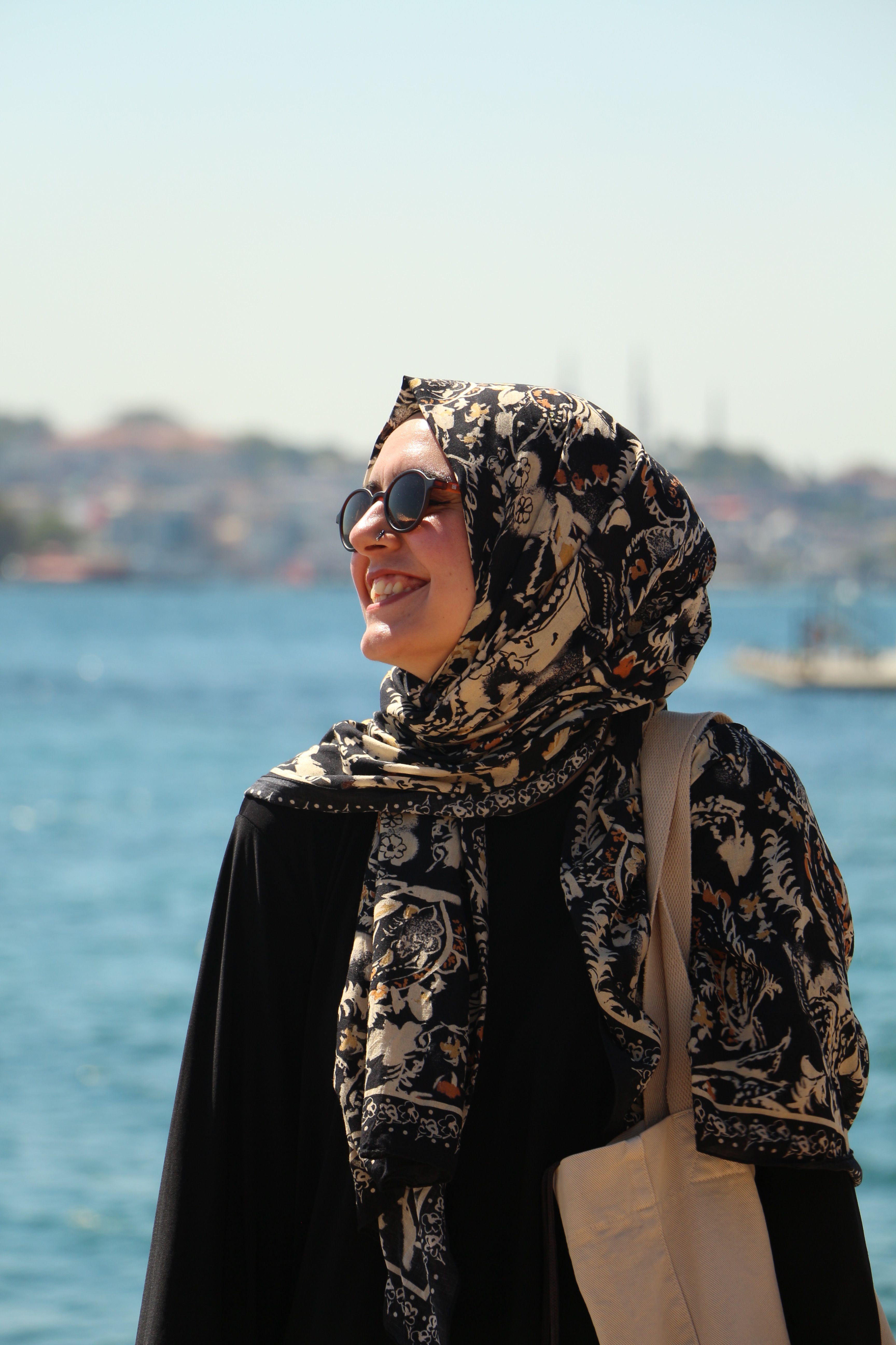 A smiling woman in hijab enjoying a sunny day by the sea with blurred cityscape in the background.