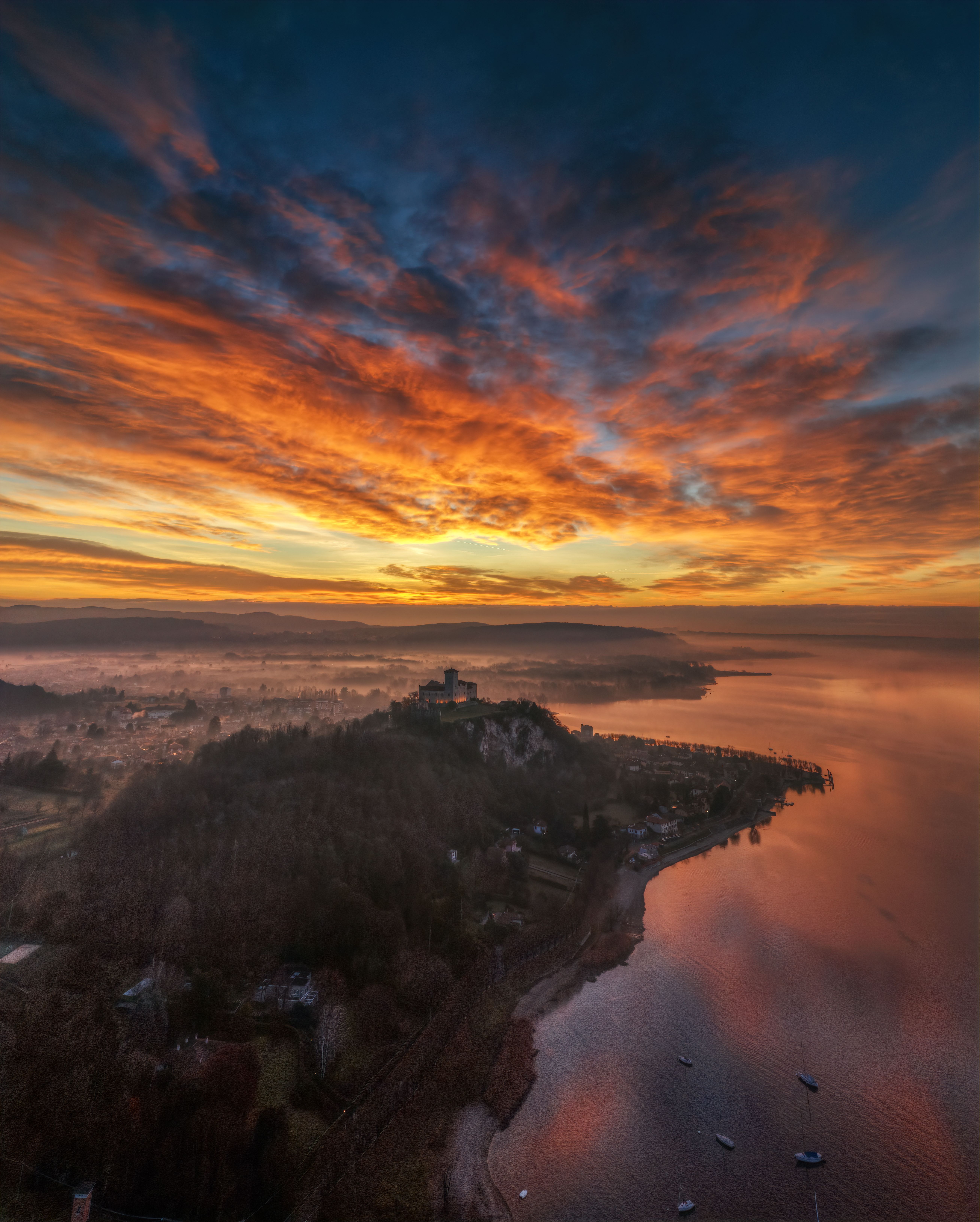 Breathtaking aerial view of a vibrant sunset over Santa Caterina del Sasso, Italy, highlighting dramatic skies and tranquil lake.