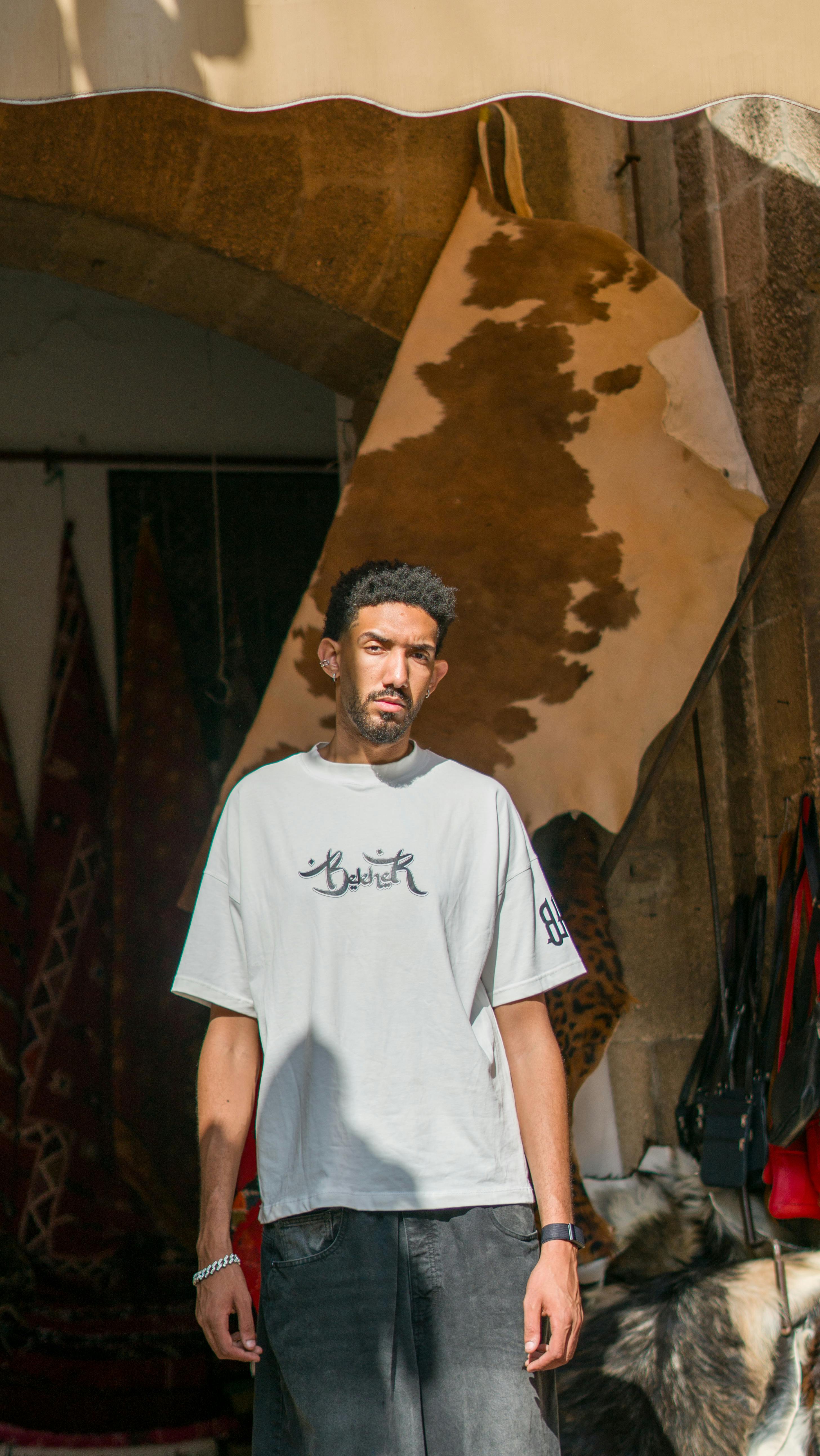 A man standing casually in a Moroccan archway surrounded by textiles, bathed in sunlight.