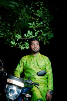A man in vibrant traditional dress sits on a motorcycle, surrounded by greenery at night.