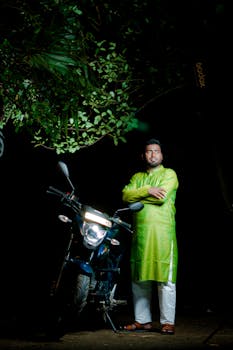 A man posing confidently beside a motorcycle under a tree at night.