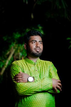 A confident man in traditional green attire poses outdoors with crossed arms. Night setting.