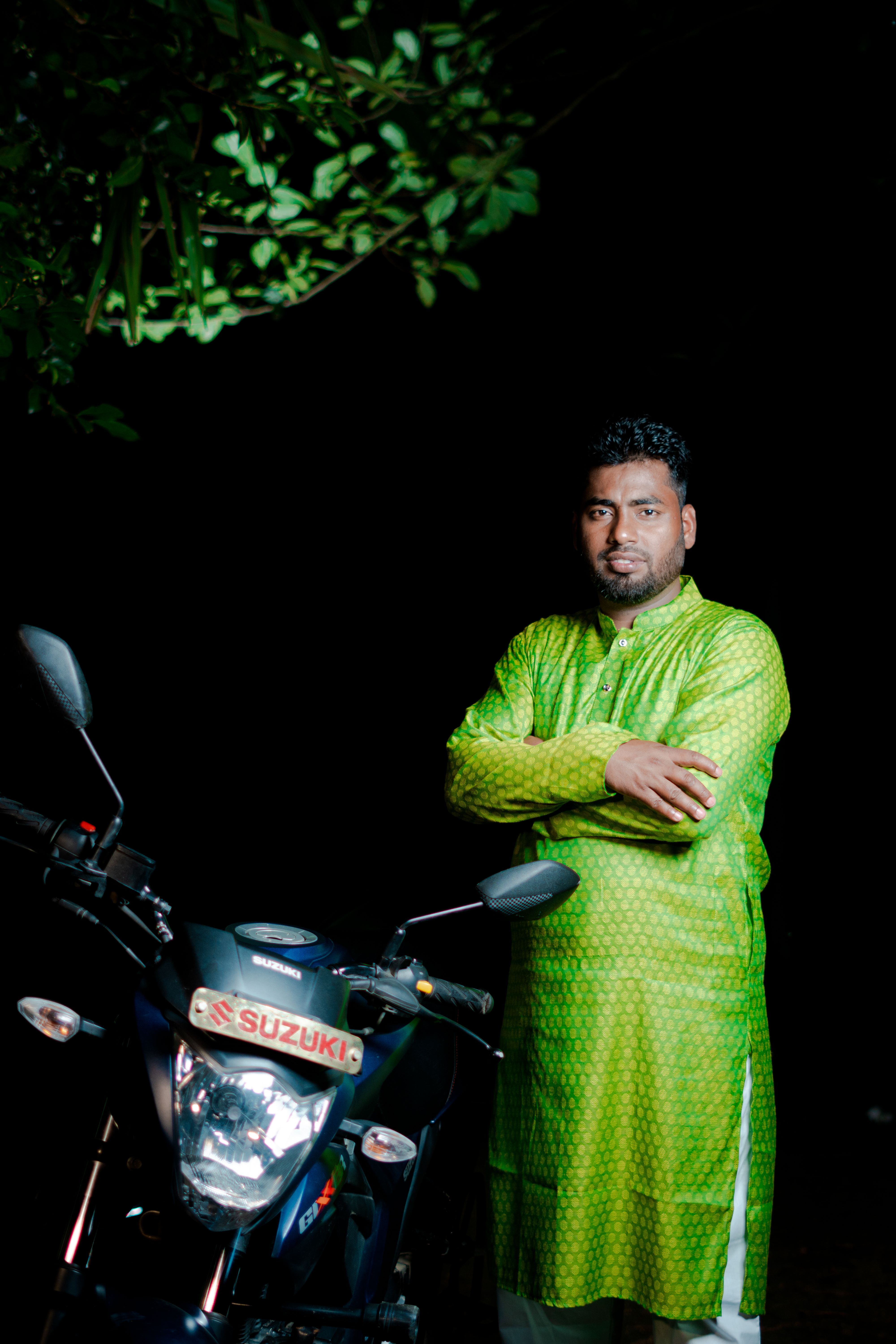 Man in green traditional attire standing beside a motorcycle at night, showcasing cultural fashion.