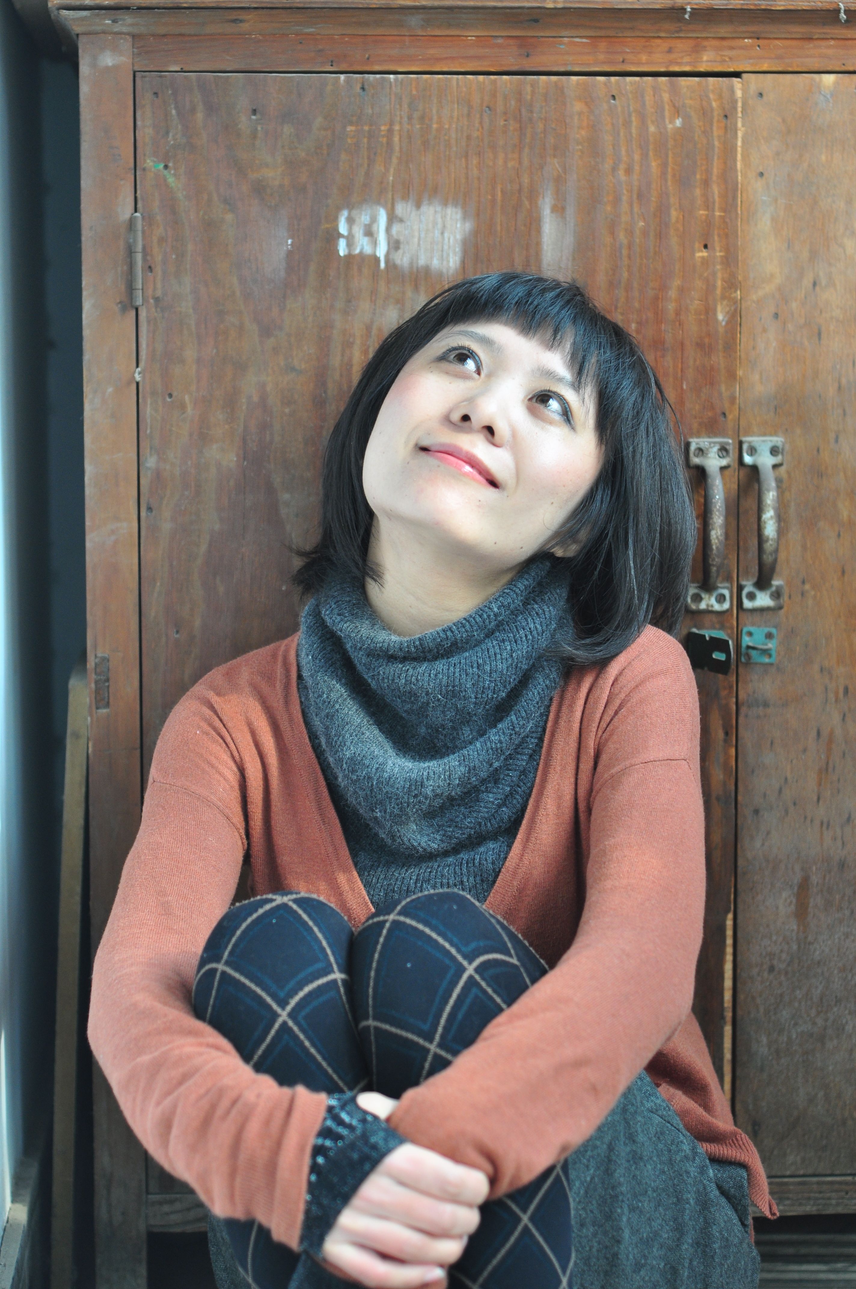 A woman in casual attire sits thoughtfully by a vintage cabinet indoors.