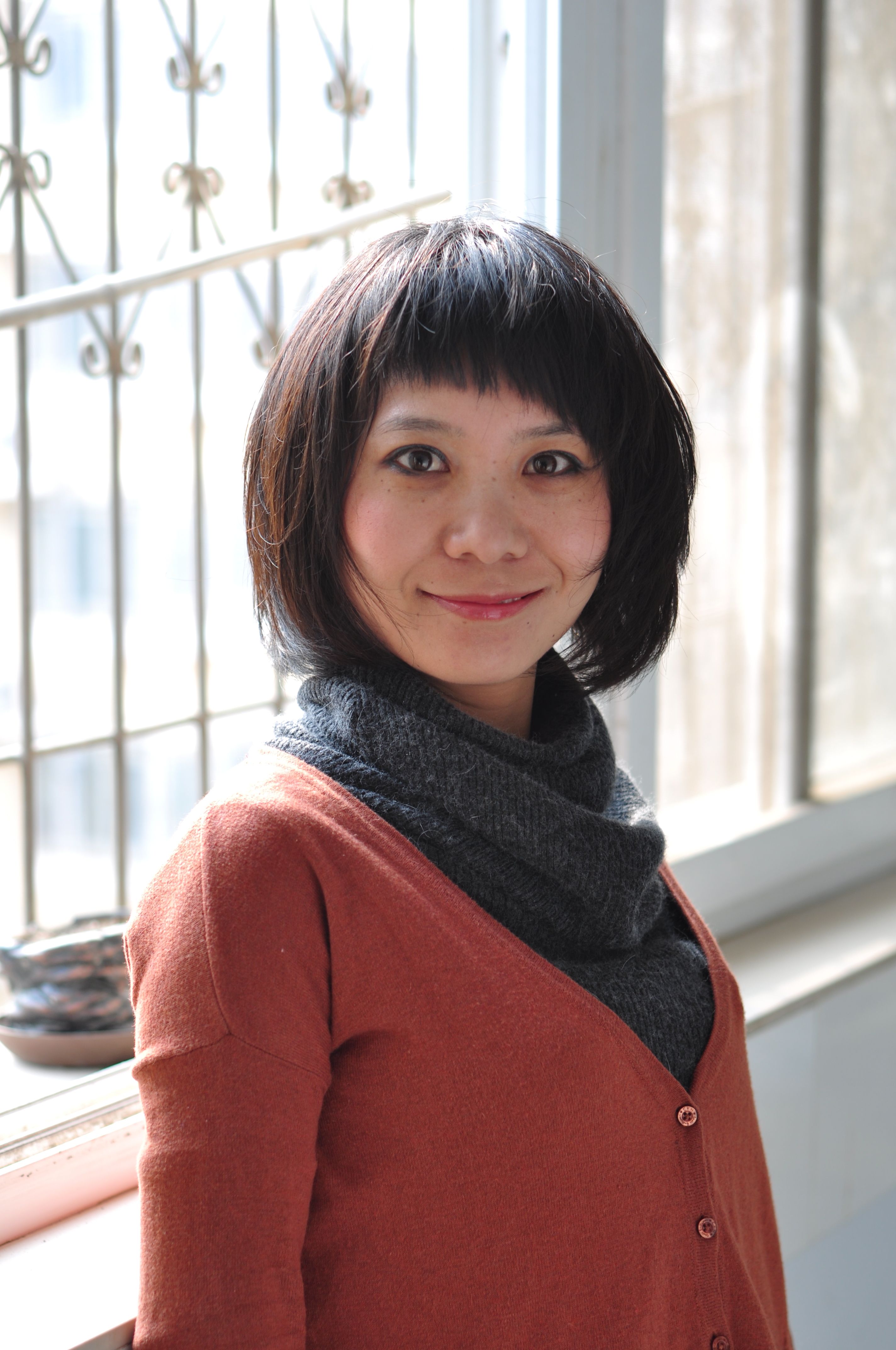 A woman in a casual outfit smiles while standing by a window, creating a warm and welcoming atmosphere.