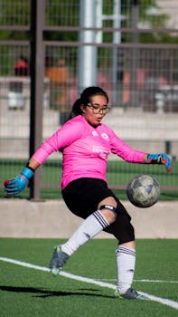 A young female goalkeeper in pink jersey making a play on a sunny soccer field.