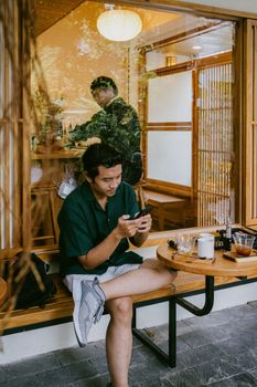 Young man using smartphone at an outdoor cafe with warm wooden decor. Perfect blend of leisure and technology.