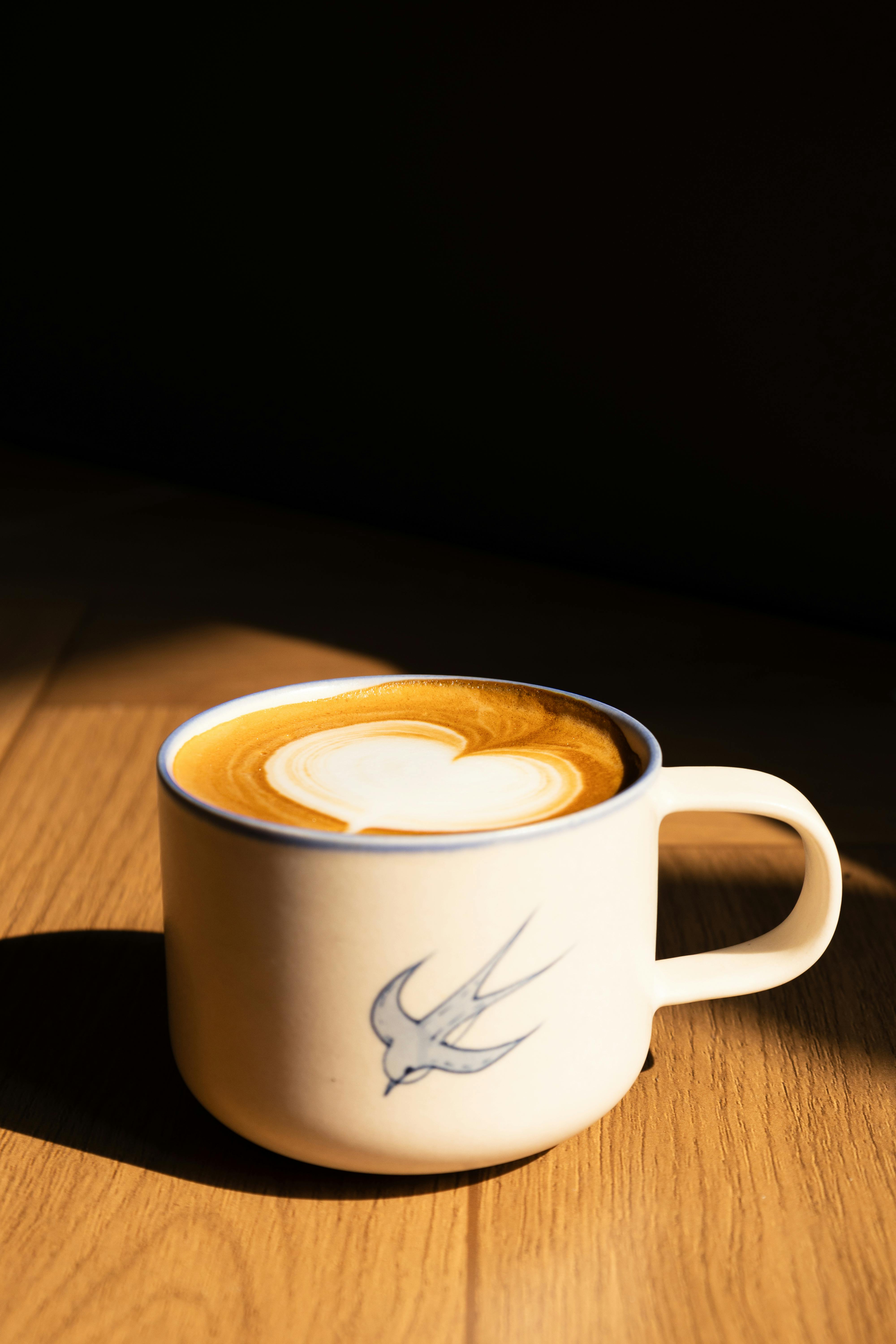 A beautifully crafted latte with heart latte art in a ceramic cup, basking in warm sunlight on a wooden surface.
