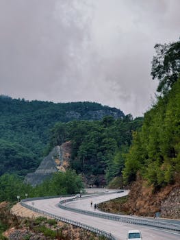 Winding mountain road with cyclists and a car in lush green forest landscape.