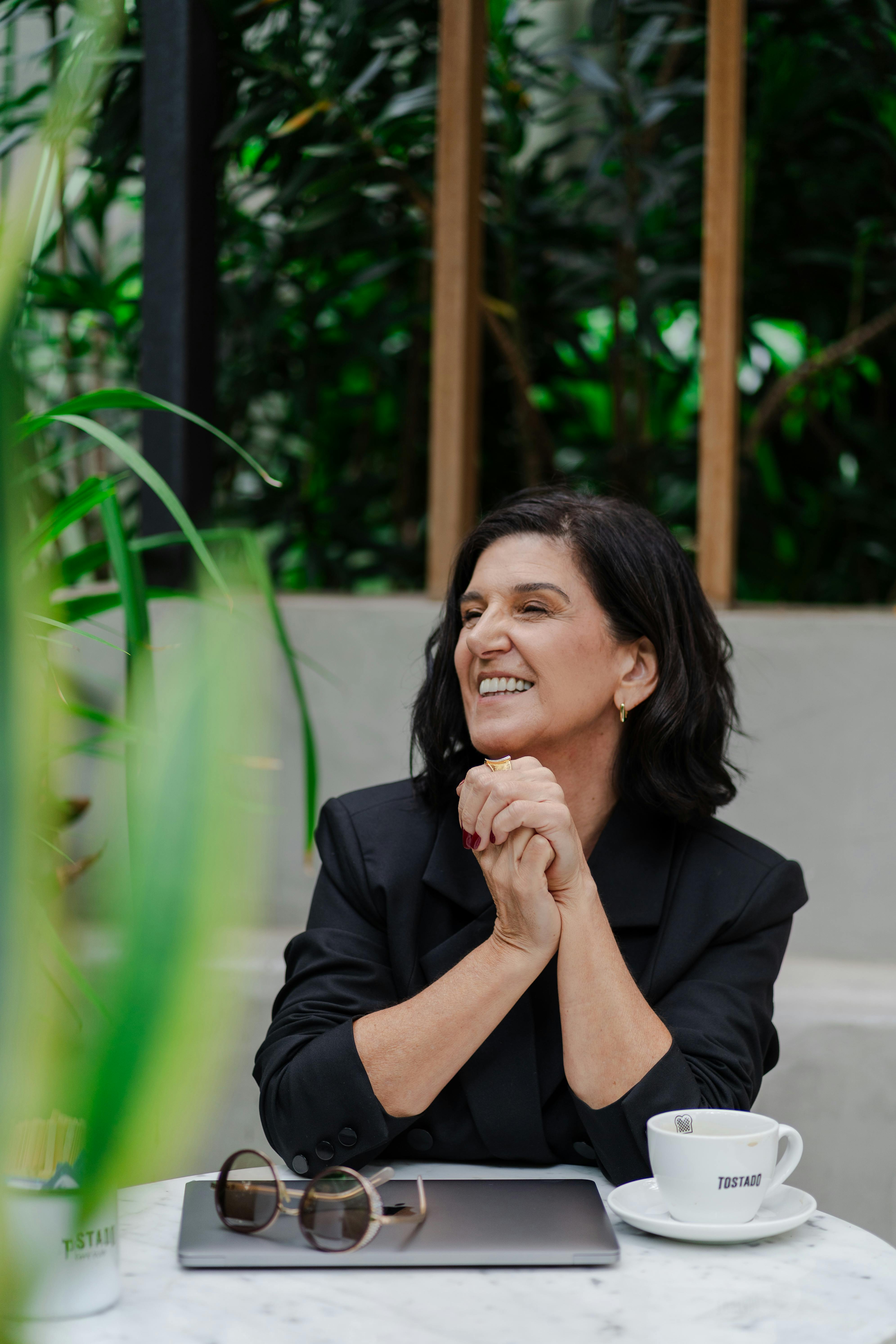 Elegant woman in black suit sitting at cafe table with laptop and coffee cup, surrounded by greenery.