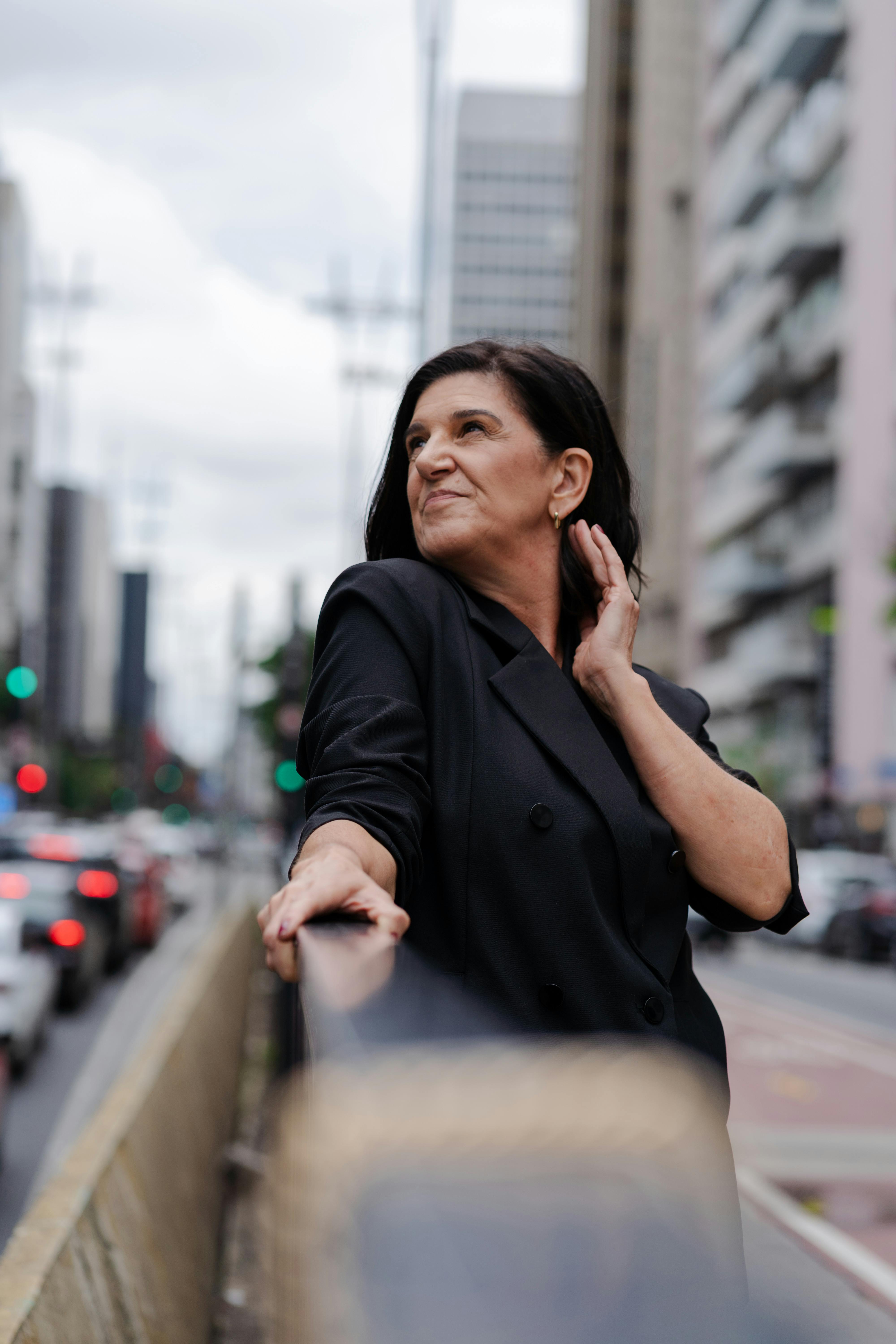Portrait of an adult woman in black coat, standing confidently on a busy city street amidst tall buildings.