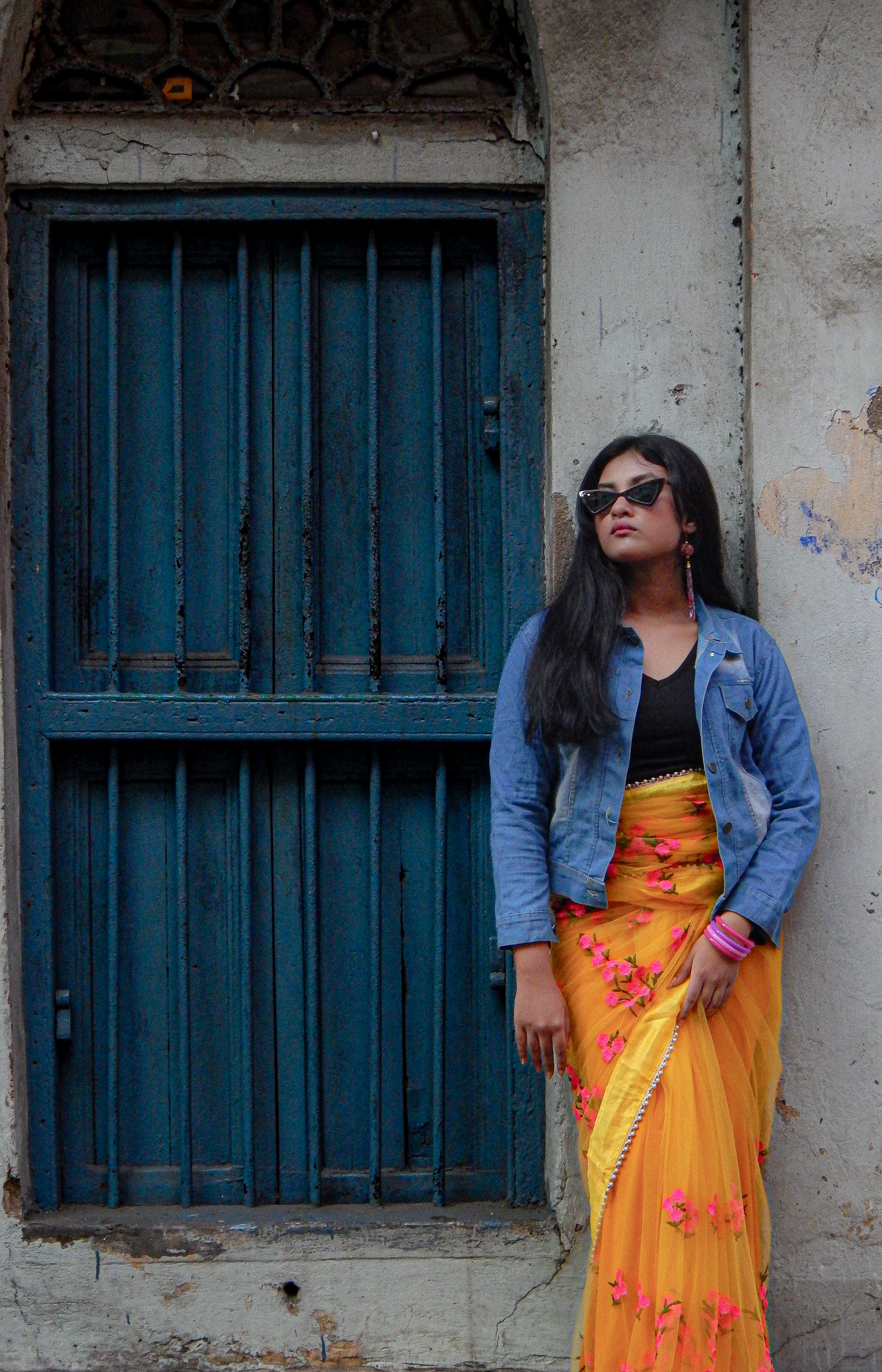 Fashionable woman in vibrant saree and denim jacket by a vintage blue door in India.
