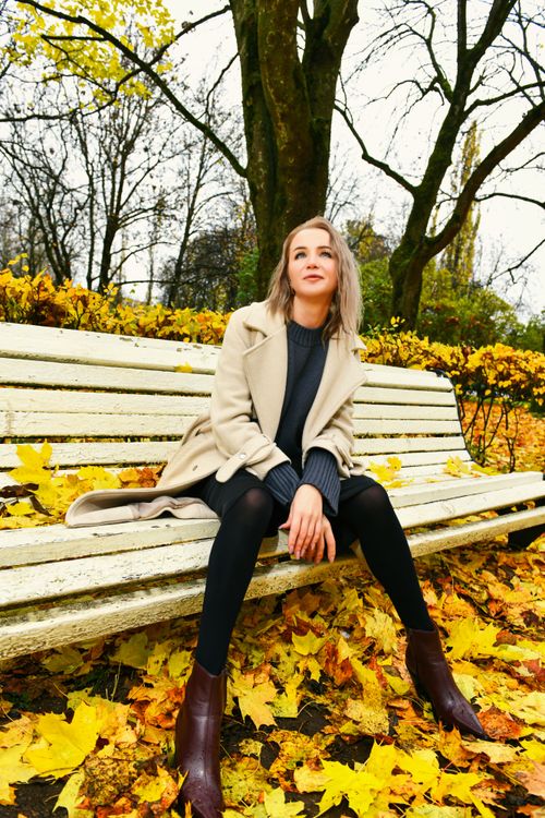 Free Young woman in fall attire sits on a park bench surrounded by vibrant autumn leaves. Stock Photo