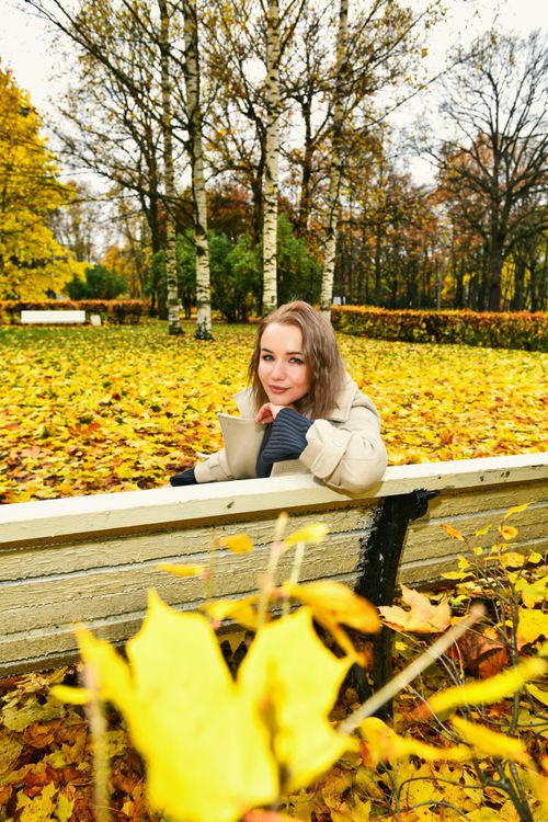 Free A woman in a cozy coat enjoys a coffee while sitting on a bench surrounded by vibrant fall foliage. Stock Photo