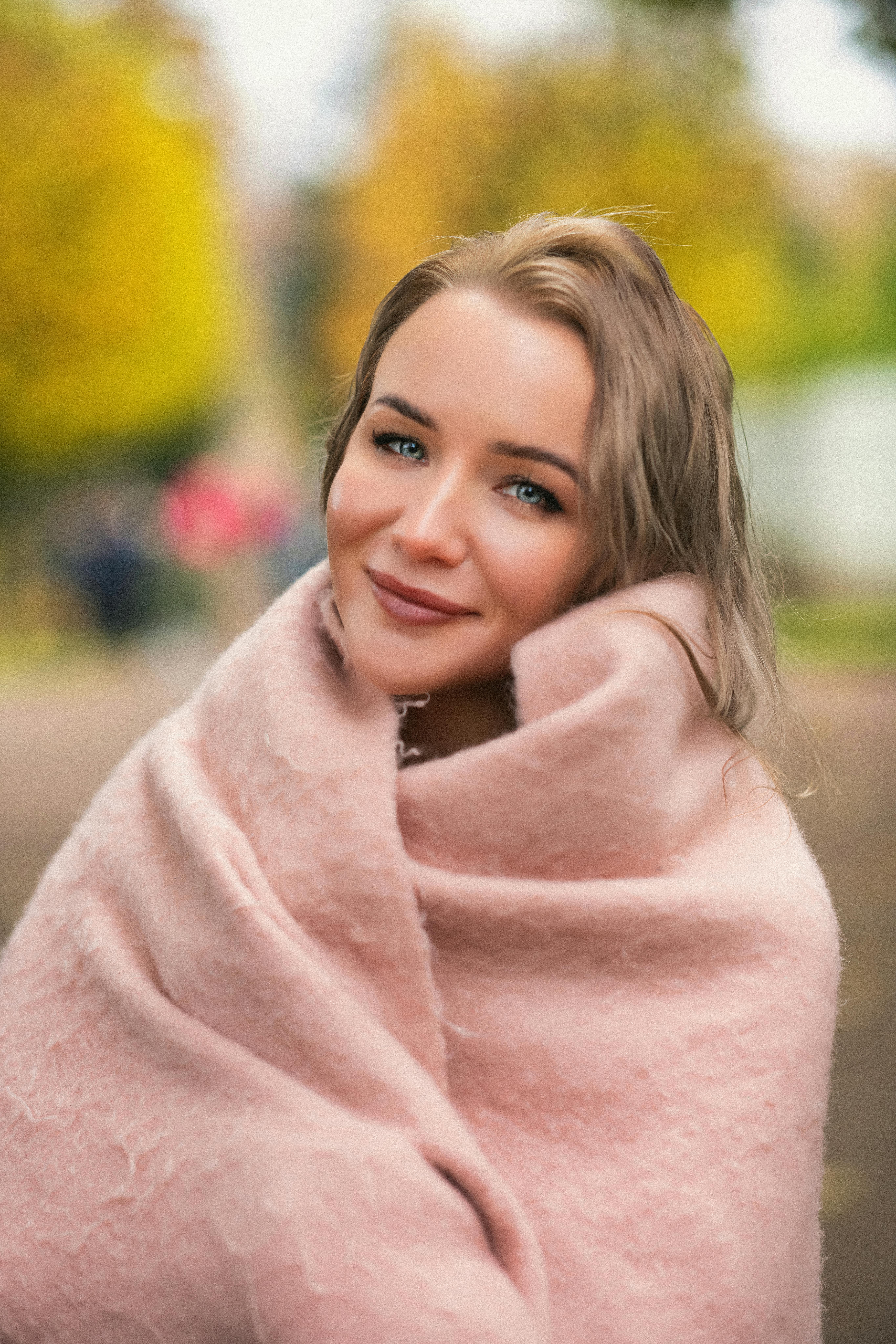 Portrait of a smiling woman wrapped in a soft pink blanket, enjoying a fall day outdoors.
