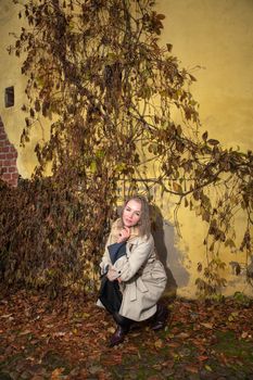 Woman in trench coat kneeling by autumn leaves on a sunny day.
