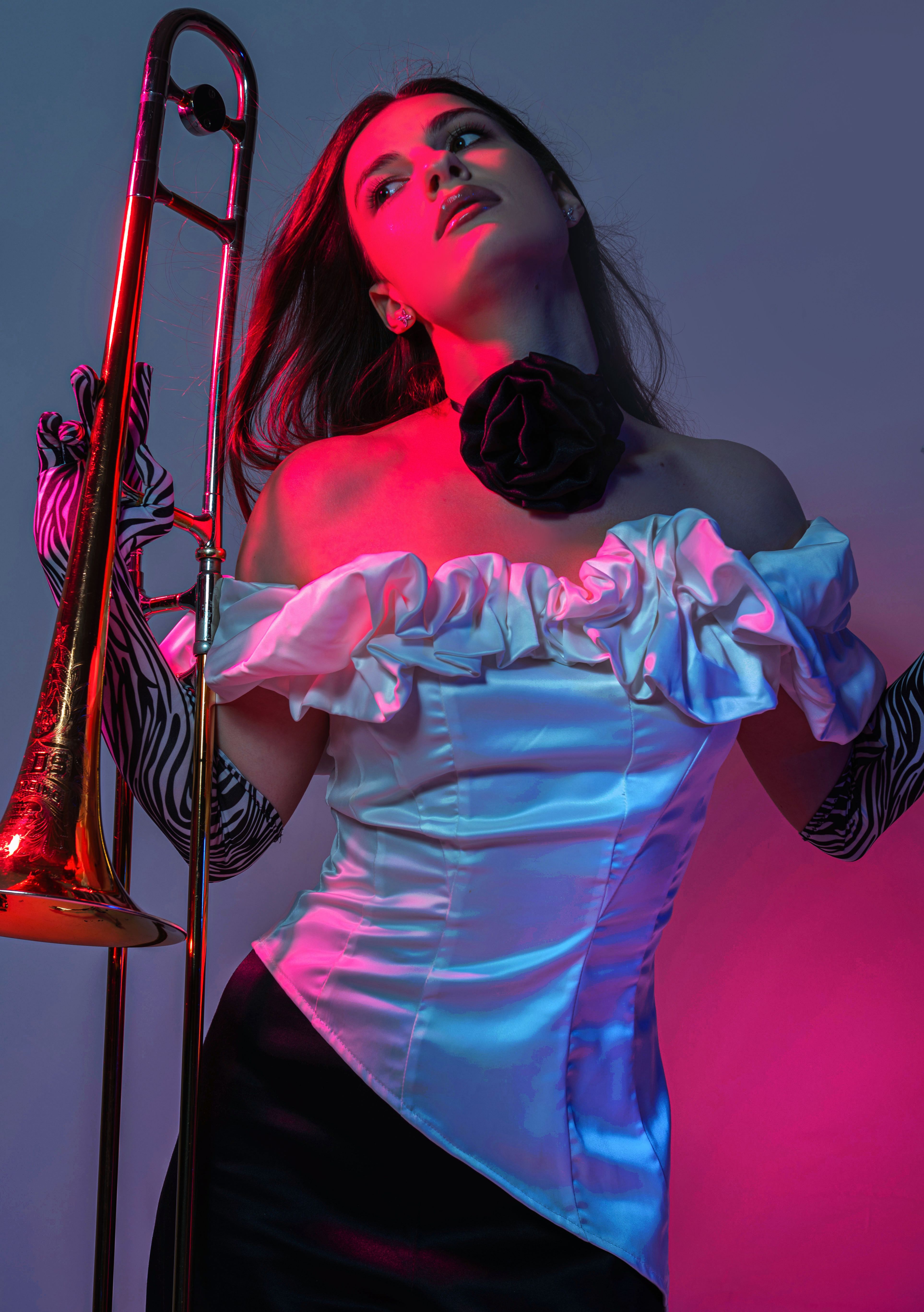 Stylish woman in elegant dress poses with trombone under vibrant neon lighting, showcasing modern fashion.