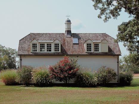A picturesque white barn with a classic weather vane framed by lush greenery and a clear sky.