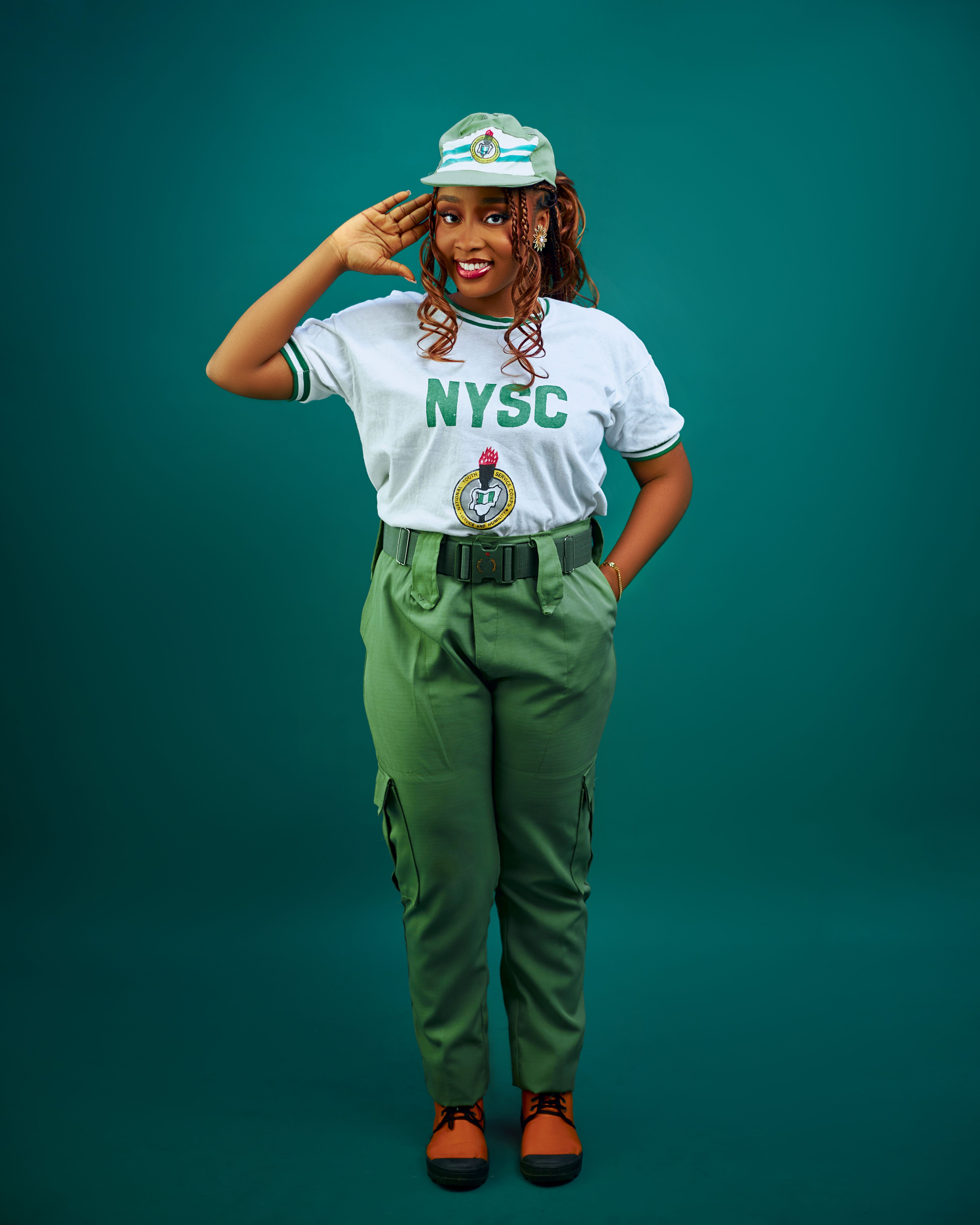 Young Woman in NYSC Uniform Saluting on Green Background · Free Stock Photo