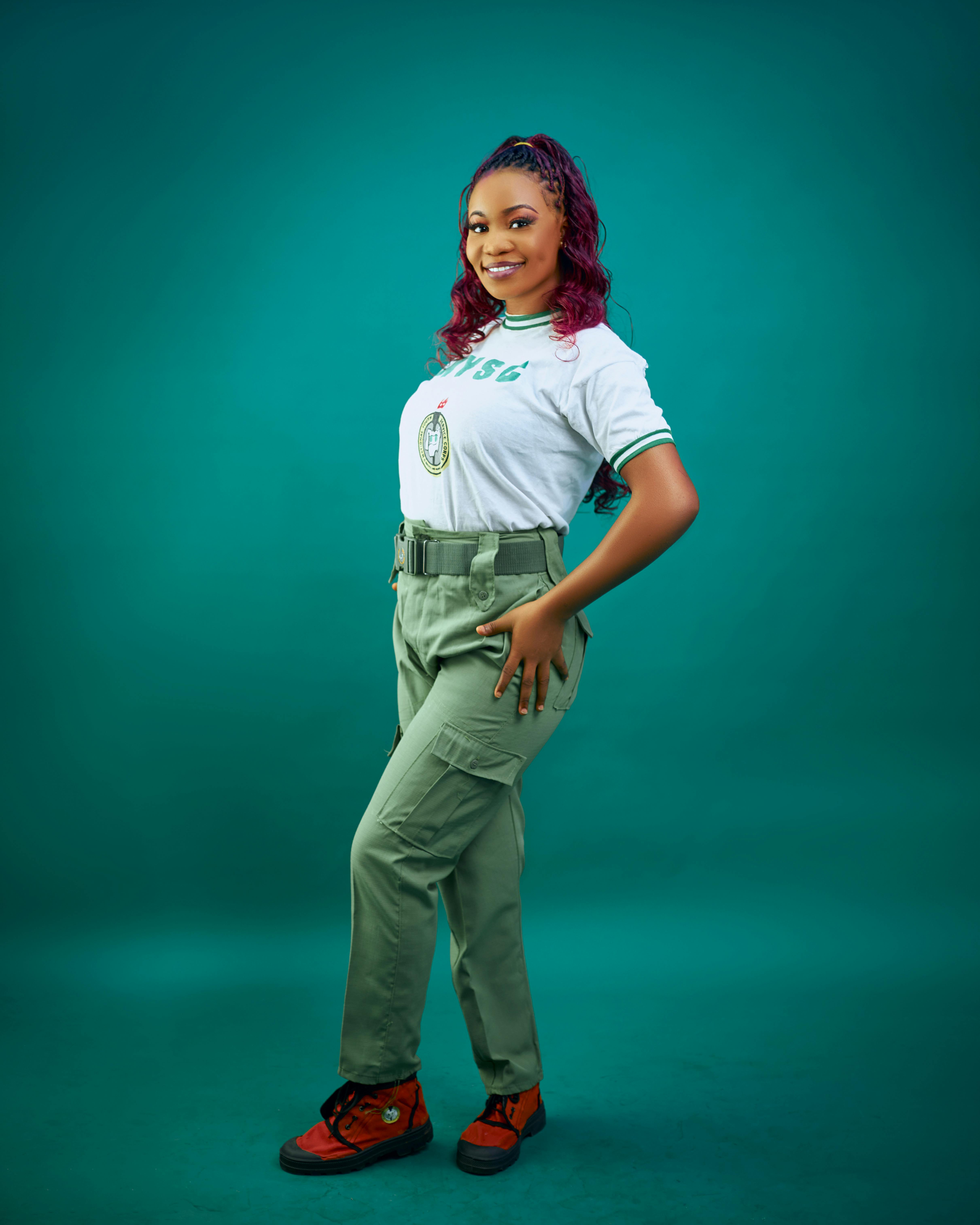 Confident young woman in green uniform posing against teal background.