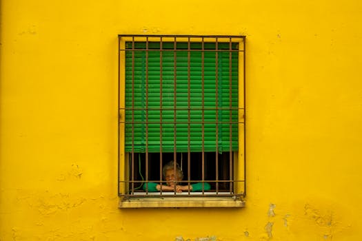 A senior woman looks out from a window with green blinds in a yellow house facade in Dozza, Italy.