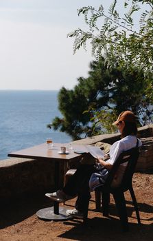 Person reading at an outdoor table by the sea, enjoying a peaceful moment.