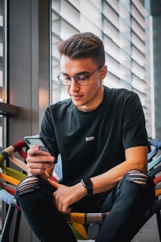 一位时尚的年轻人戴着眼镜，在室内舒适地坐着使用智能手机