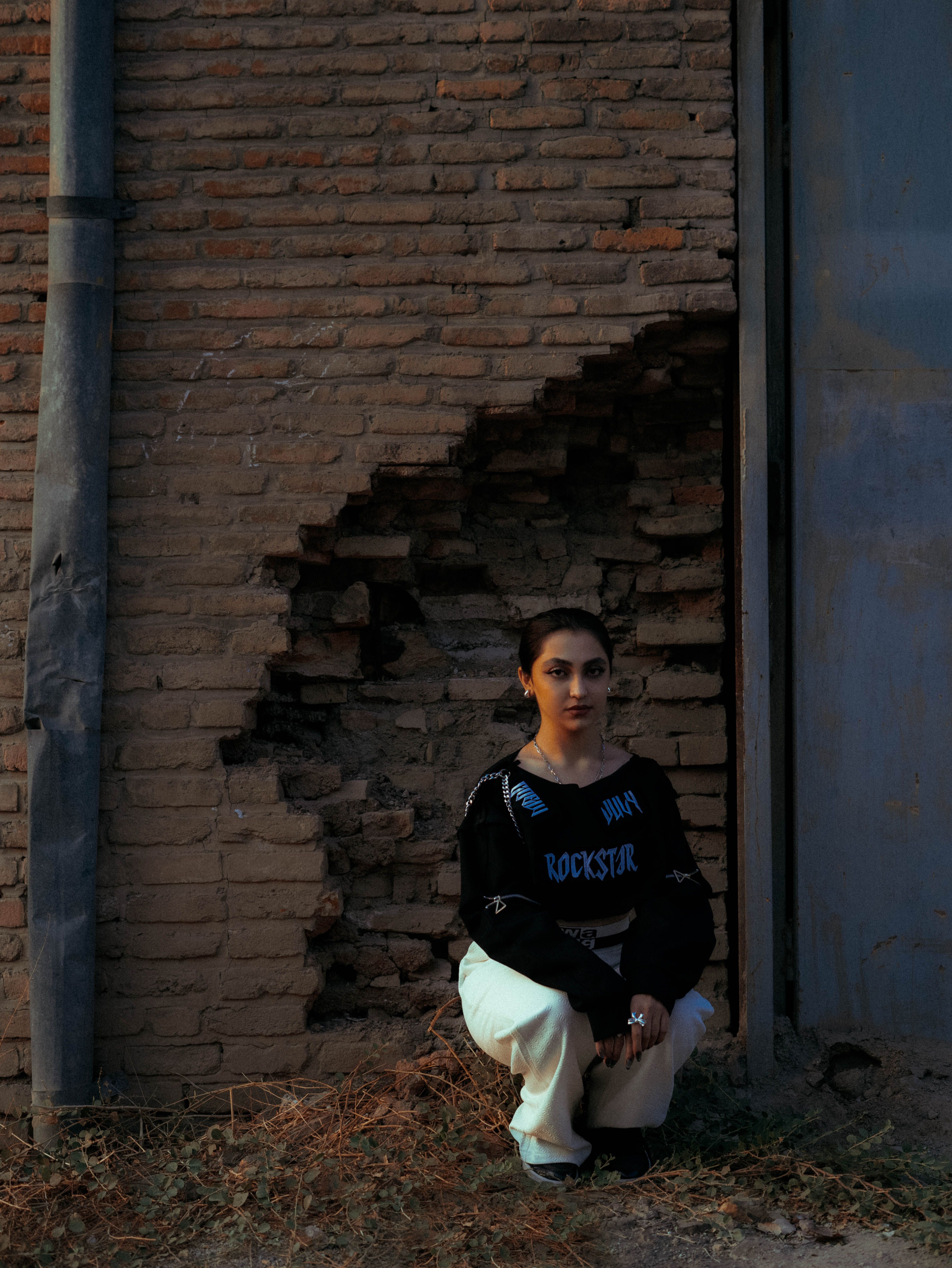 A woman crouches thoughtfully against a vintage brick wall under soft evening light.