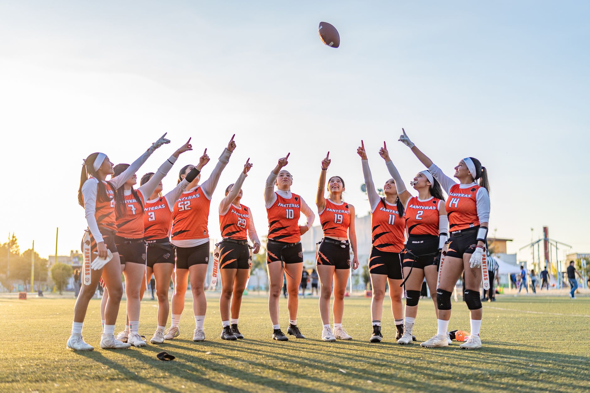 A women's flag football team raises their fingers in celebration on a sunny day.