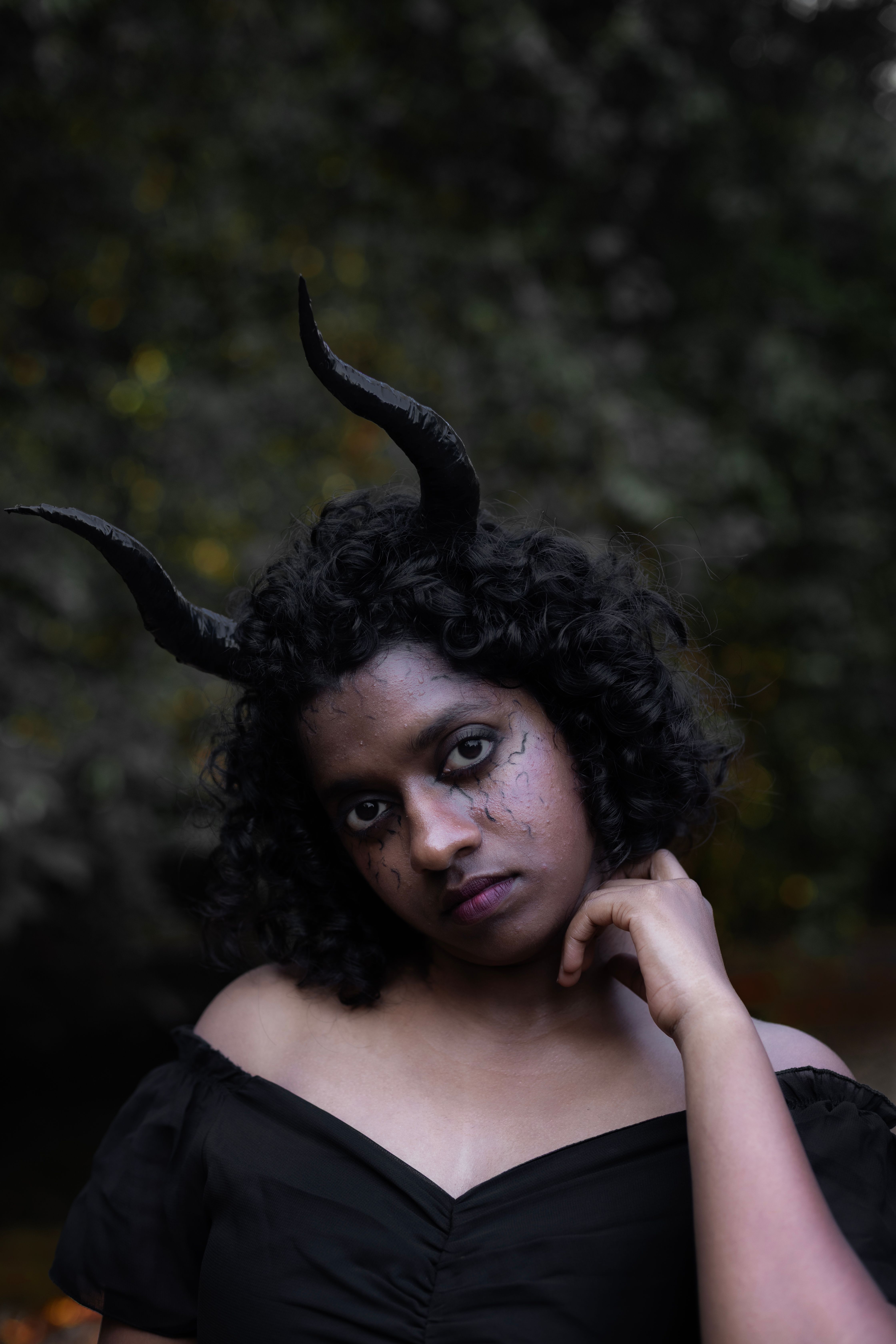 A moody, dark portrait of a person with curved horns in a forest setting.