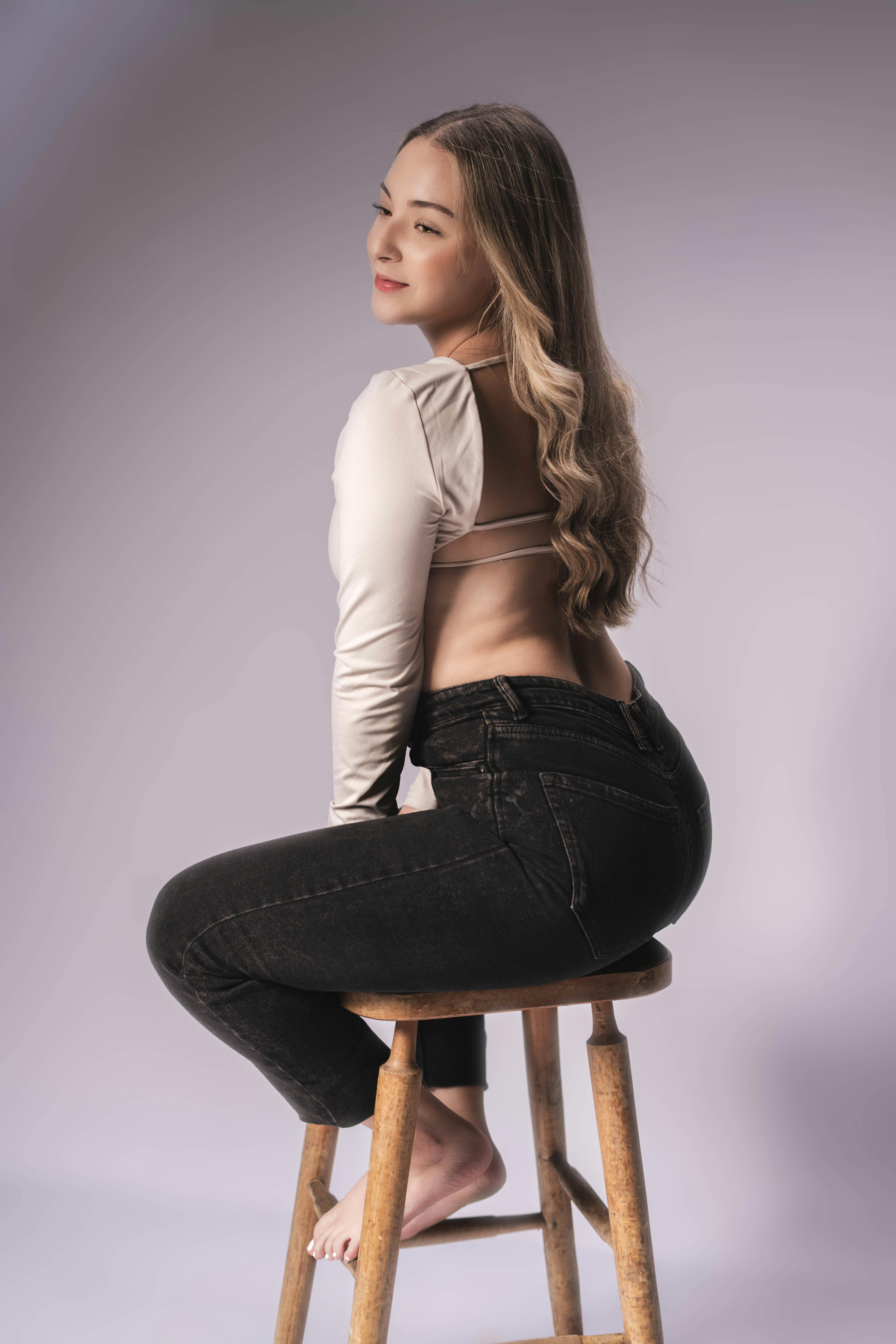 Gratis Una mujer con atuendo informal se sienta con gracia en un taburete de madera, resaltando su estilo y elegancia. Foto de stock