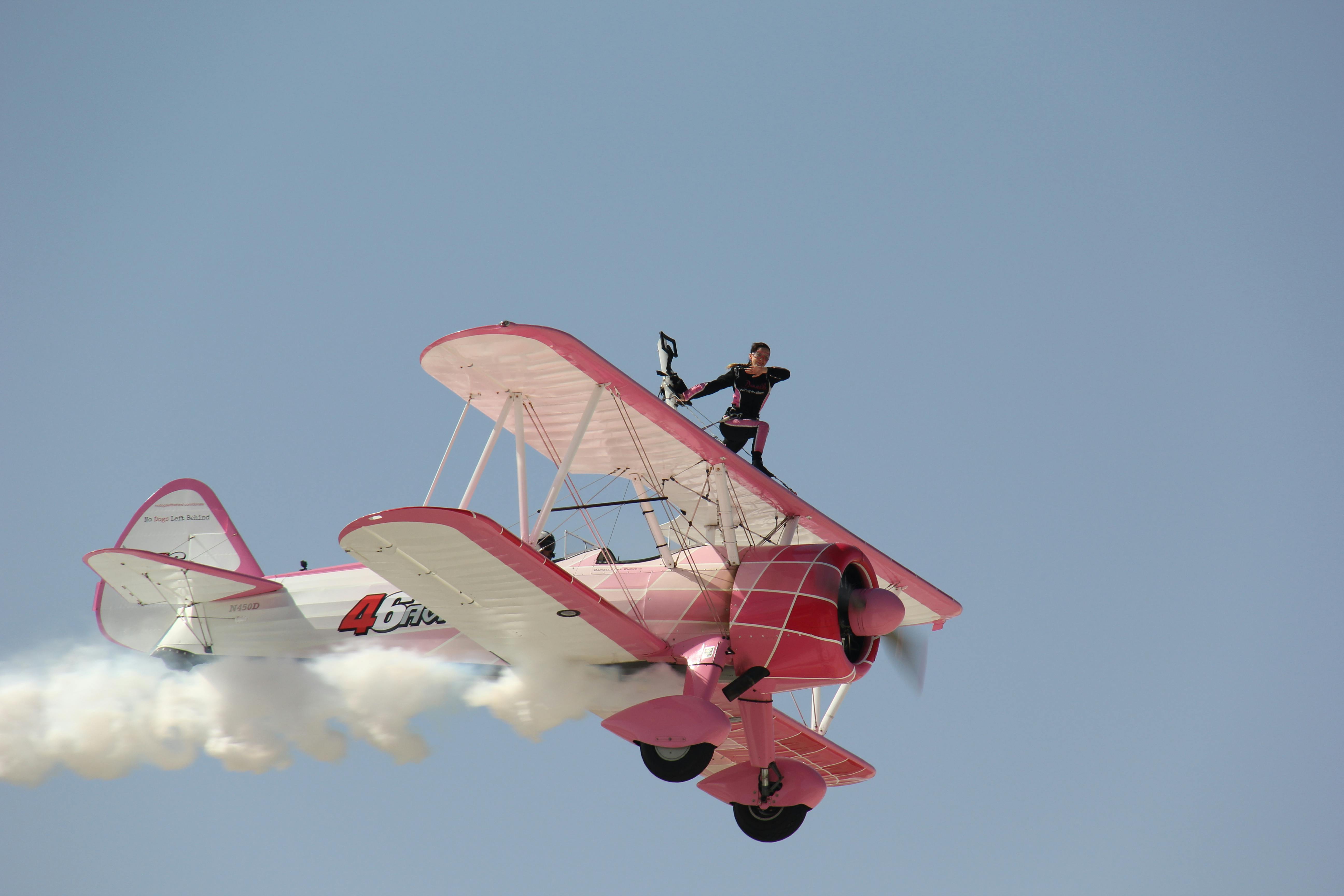 A daring wingwalker performs a stunt on a pink vintage biplane in clear sky.