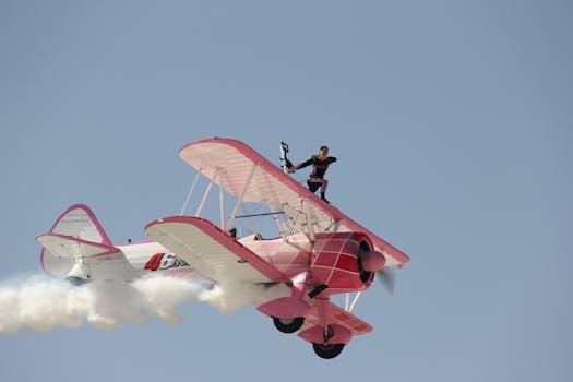 A daring wingwalker performs a stunt on a pink vintage biplane in clear sky.