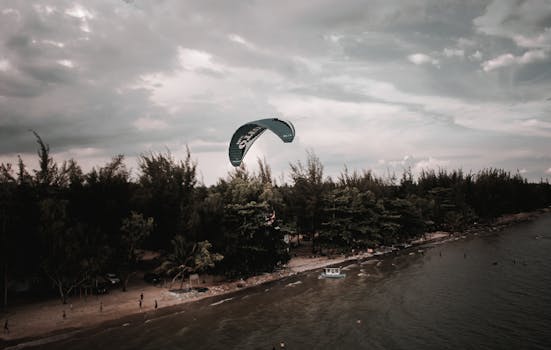 A serene aerial view of the beach with a paraglider soaring above, set against a cloudy sky.
