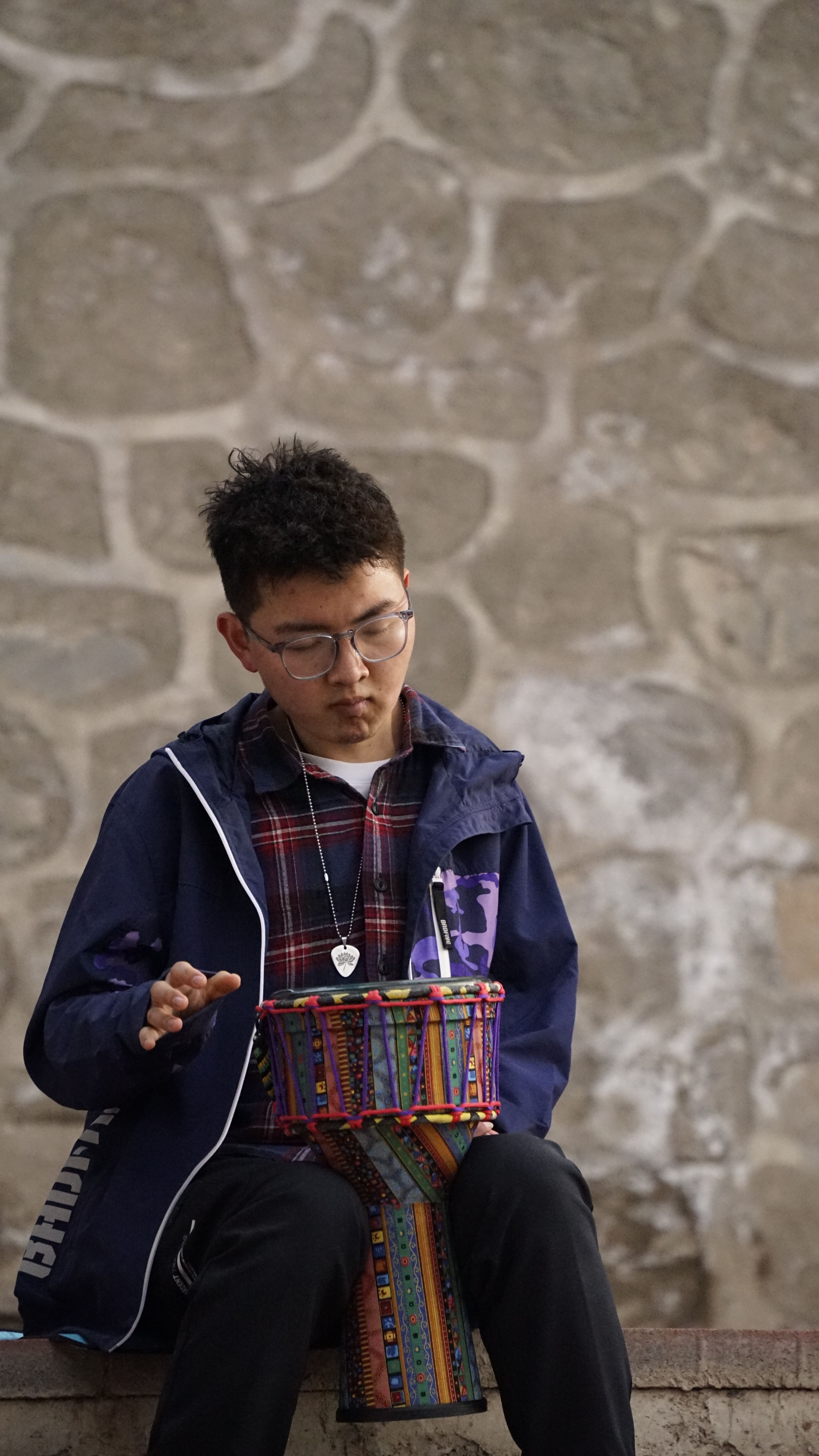 A young man focuses on playing a bright, colorful djembe drum against a stone wall background.