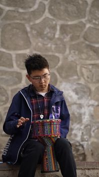 A young man focuses on playing a bright, colorful djembe drum against a stone wall background.
