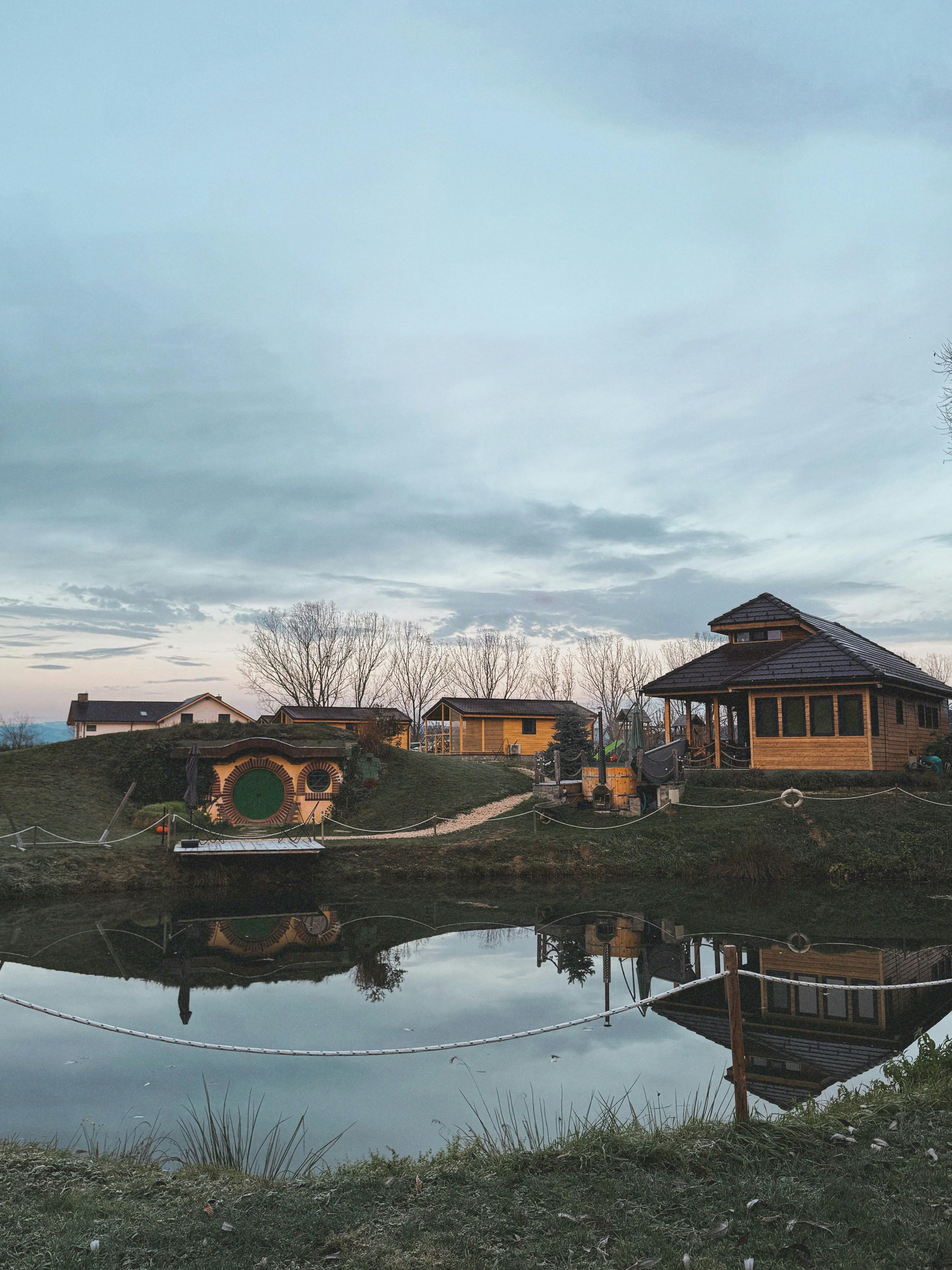 grátis Paisagem encantadora de casas rústicas, inspiradas nos hobbits, refletida em um lago tranquilo ao entardecer. Foto profissional