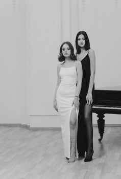 Two women in elegant dresses posing by a grand piano in a monochrome setting.