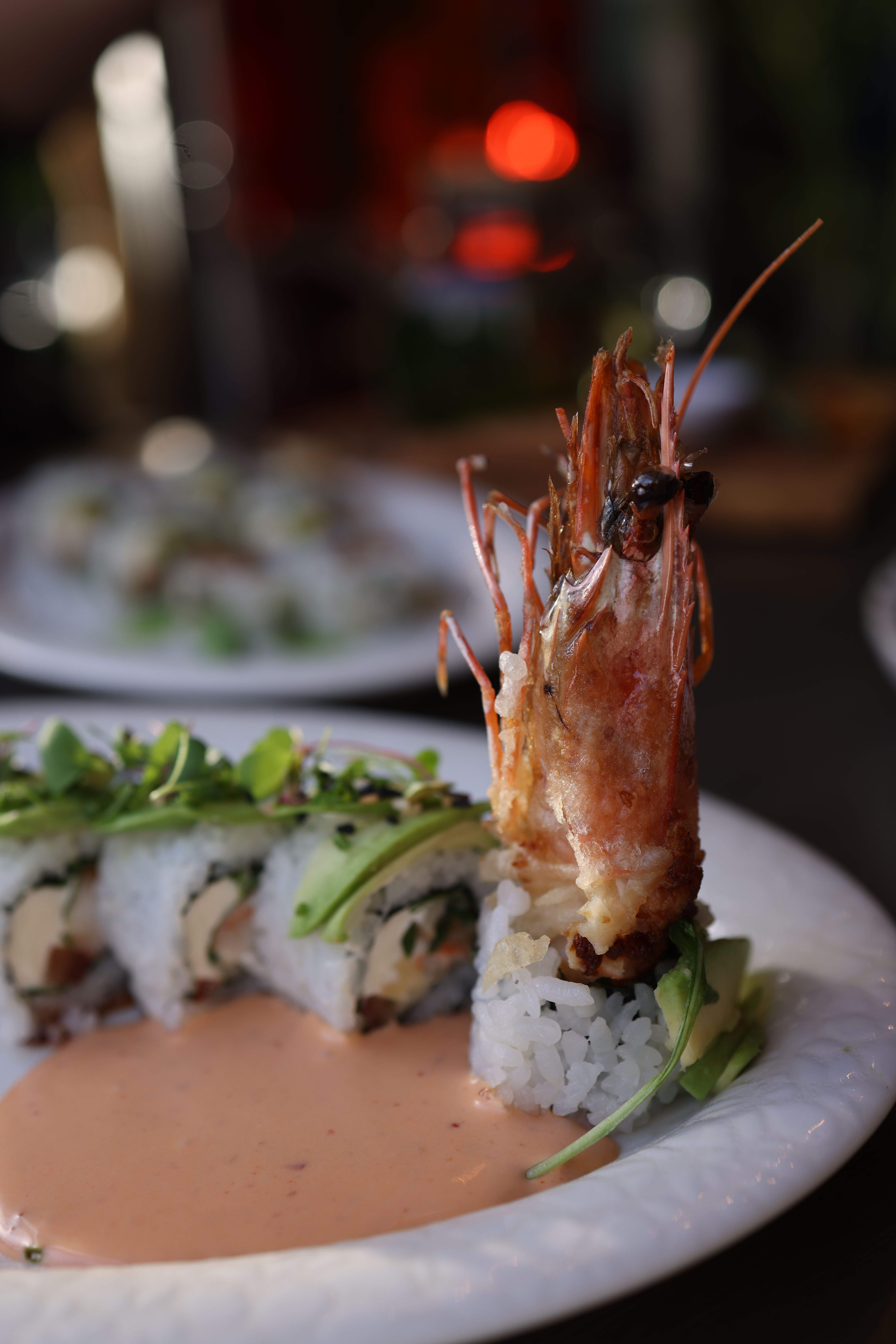 Sliced shrimp tempura sushi rolls arranged on a black plate, showing the crispy fried shrimp tail, avocado, and cucumber filling, drizzled with spicy mayo and eel sauce and topped with sesame seeds.
