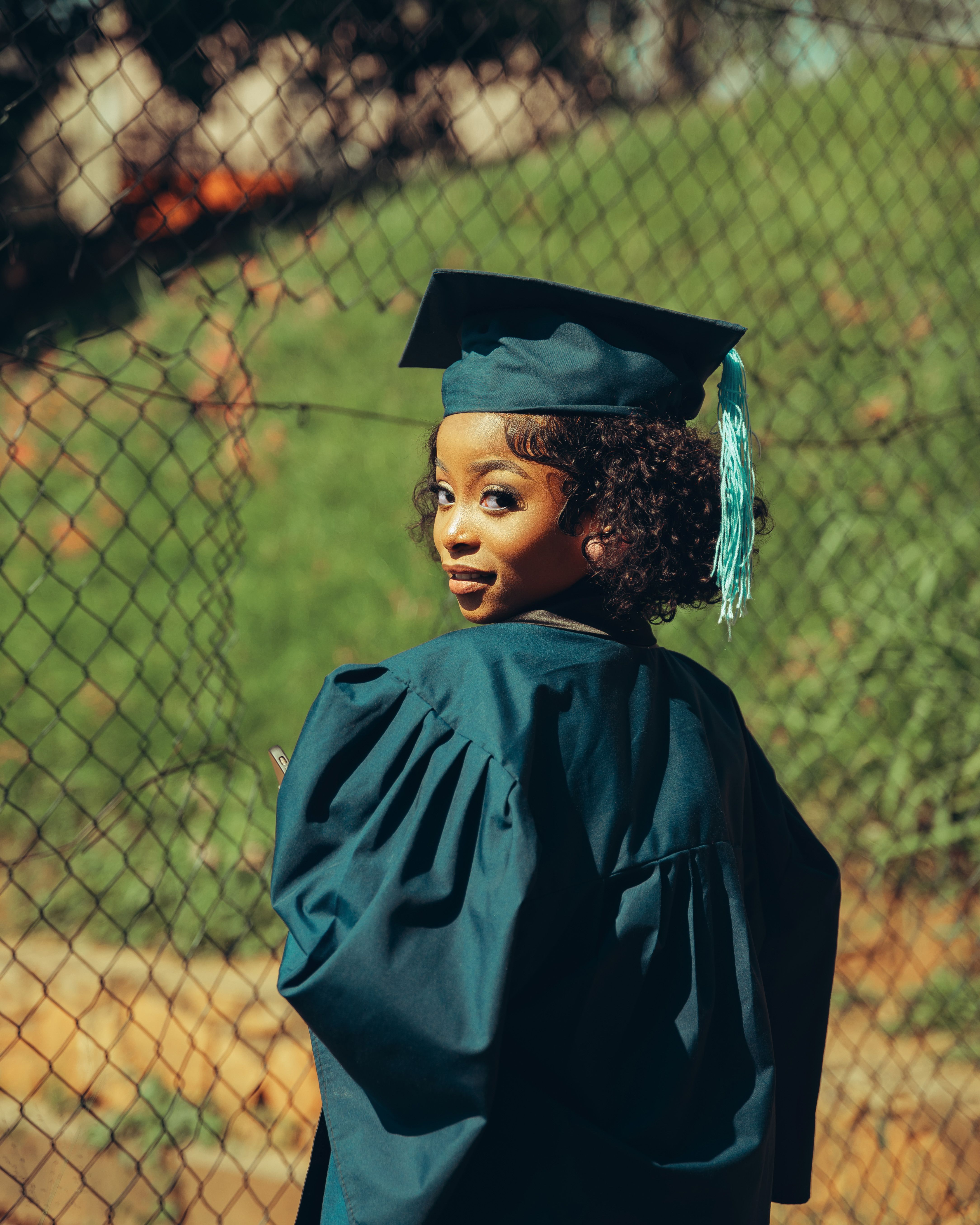 A young graduate wearing a cap and gown smiles, standing outdoors by a fence.