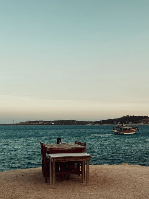 Free Empty tables by the sea with a distant boat at sunset in Taşucu, Mersin, Türkiye. Stock Photo