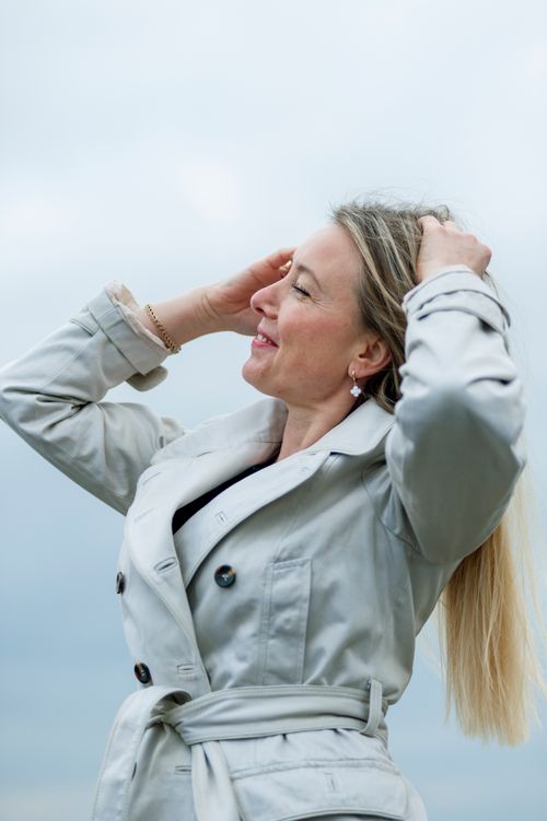 Free A woman in a light gray trench coat smiling and enjoying the breeze on a cloudy day. Stock Photo