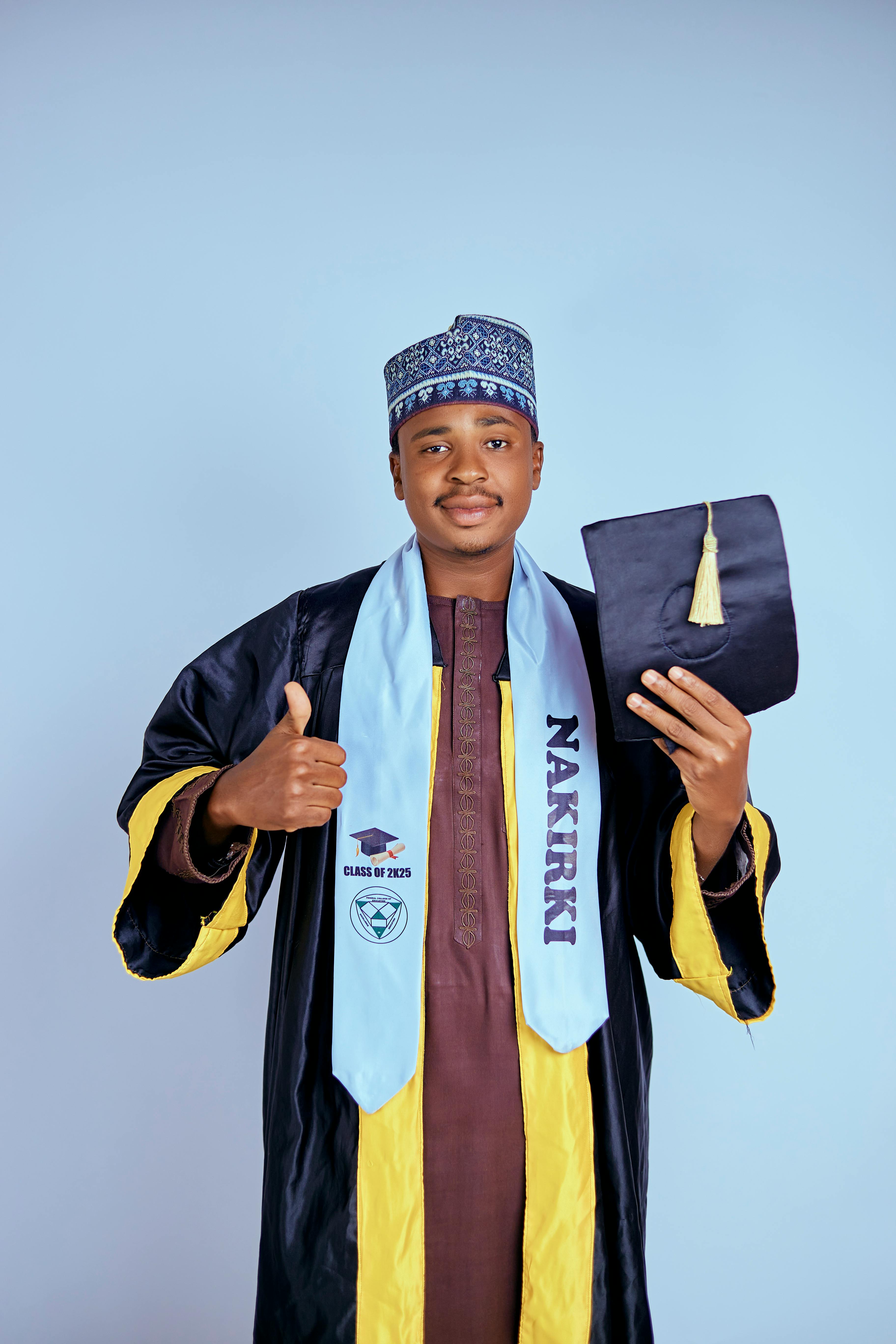 Portrait of a young graduate in traditional attire holding diploma and giving thumbs up.