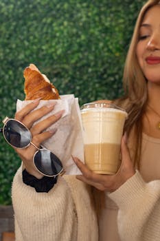 Blonde woman holding a croissant and iced latte, showcasing a chic, relaxed cafe vibe.