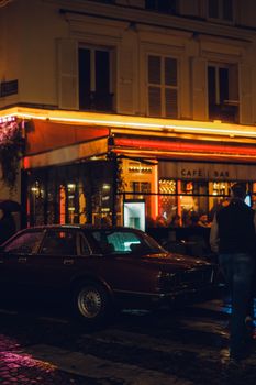 A vintage car parked outside a warmly lit Parisian cafe bar on a rainy night.