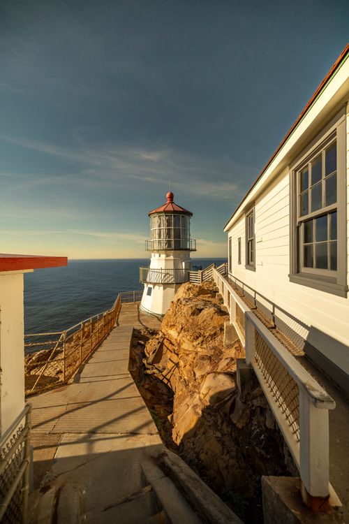 Free A picturesque lighthouse perched on a rocky coastline against a clear blue sky, ideal for travel and maritime themes. Stock Photo