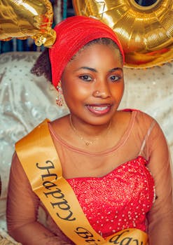 Woman in red dress and headscarf celebrating birthday indoors with balloons.