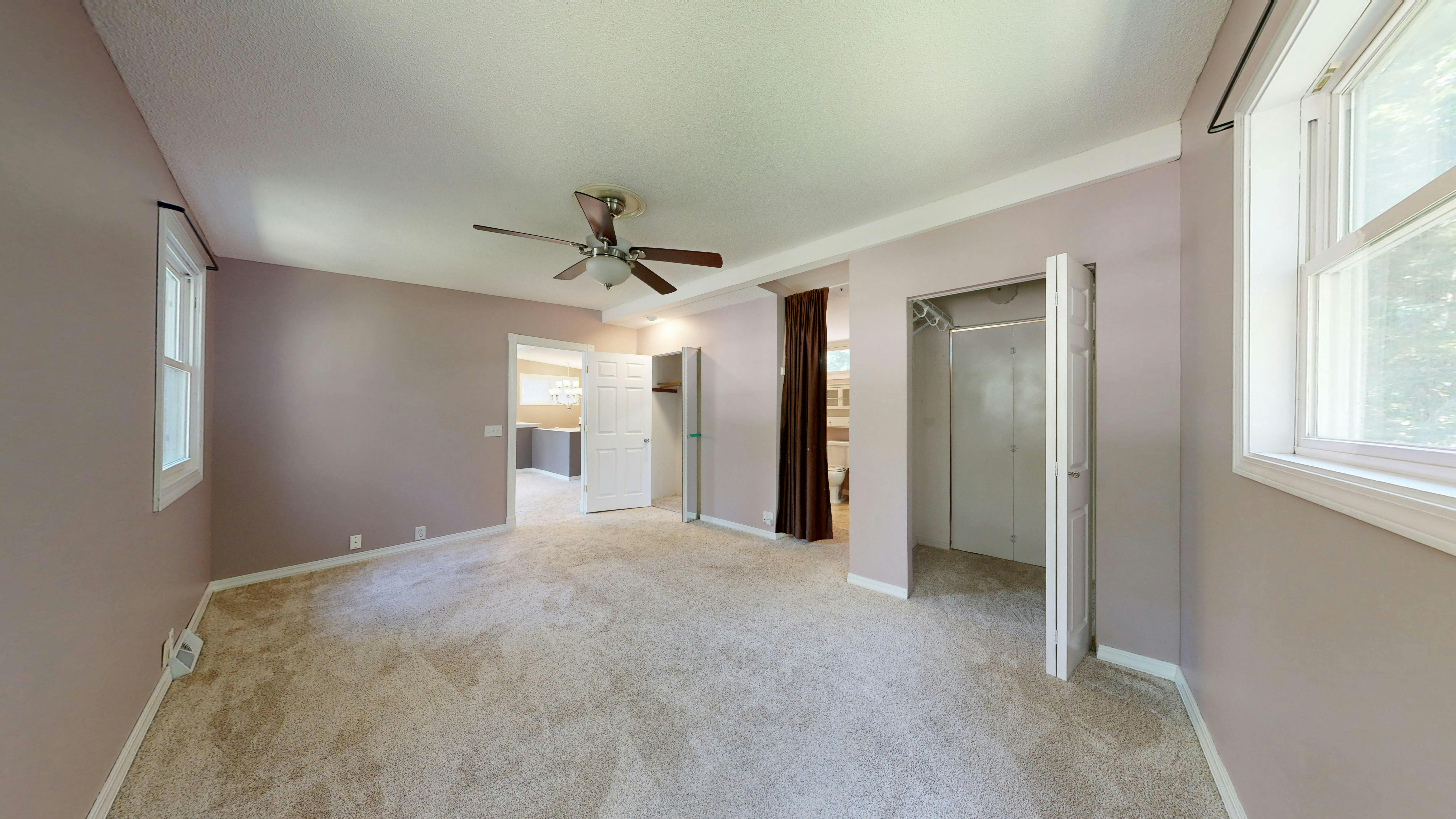 Bedroom with dusty rose walls, beige carpet, doorways to bathroom and other rooms, and a ceiling fan.